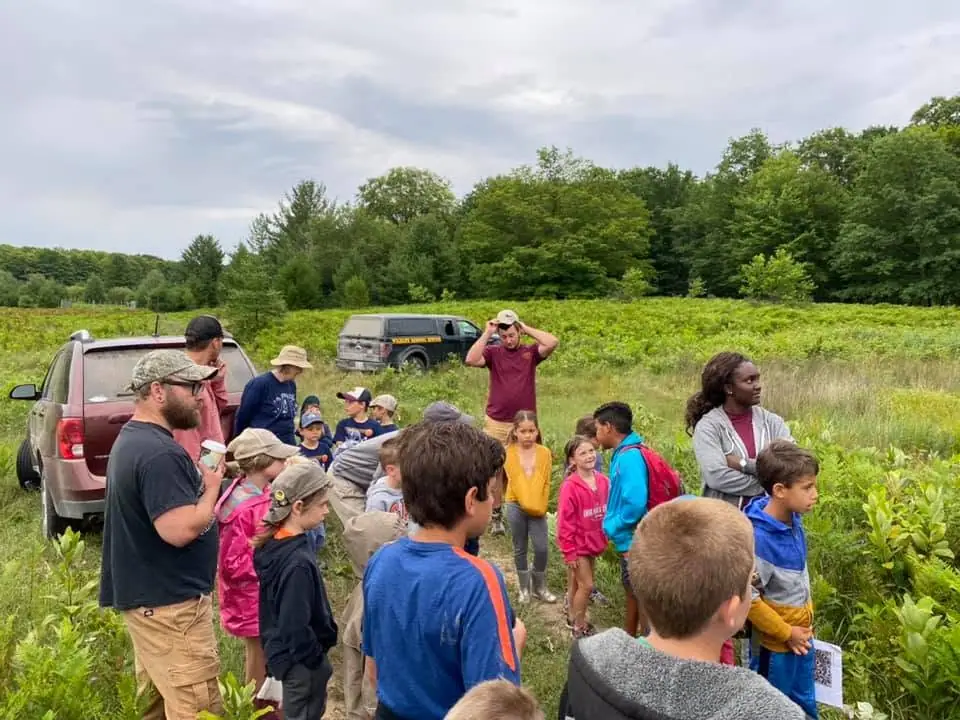 Adults and children gather beside vehicles in a grassy field bordered by trees.