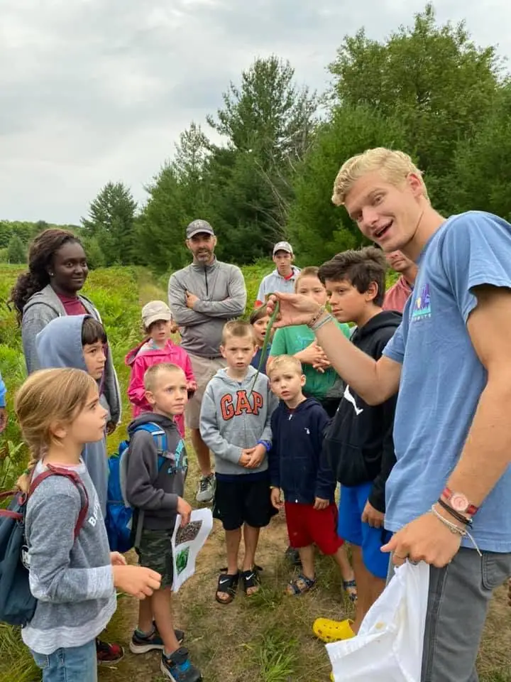 Children and adults gather outdoors as a man holds up a long green plant stem.
