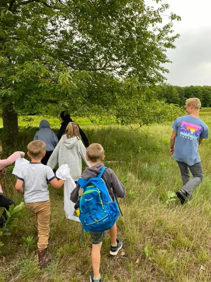 Children and adults walk through tall grass beneath a tree, carrying white bags and wearing backpacks.
