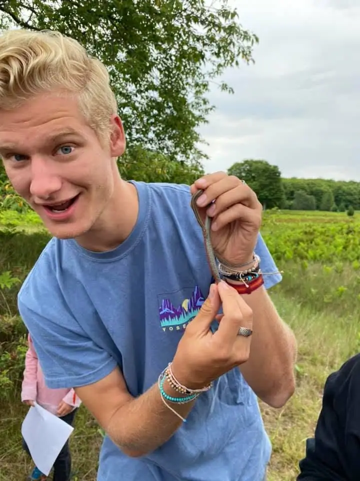 A smiling young man holds a small snake outdoors, with fields and trees behind him.