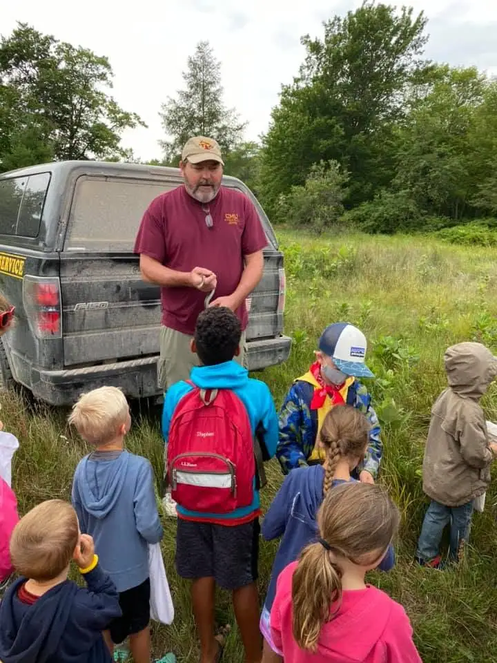 An adult stands with a group of children beside a pickup truck in a grassy clearing.