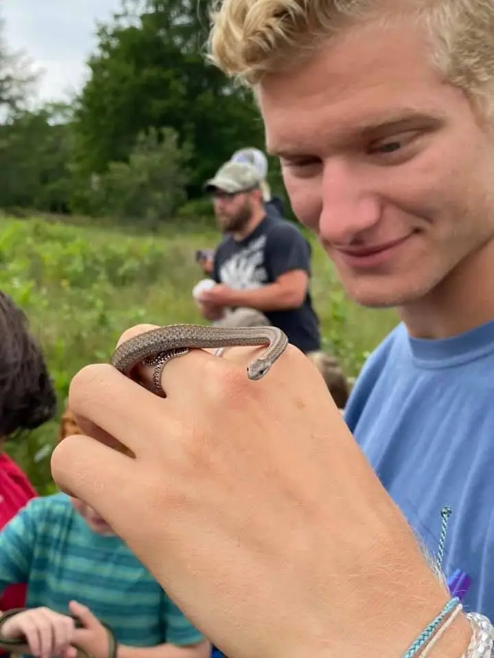 A smiling young man holds a slender snake looped around his hand outdoors.