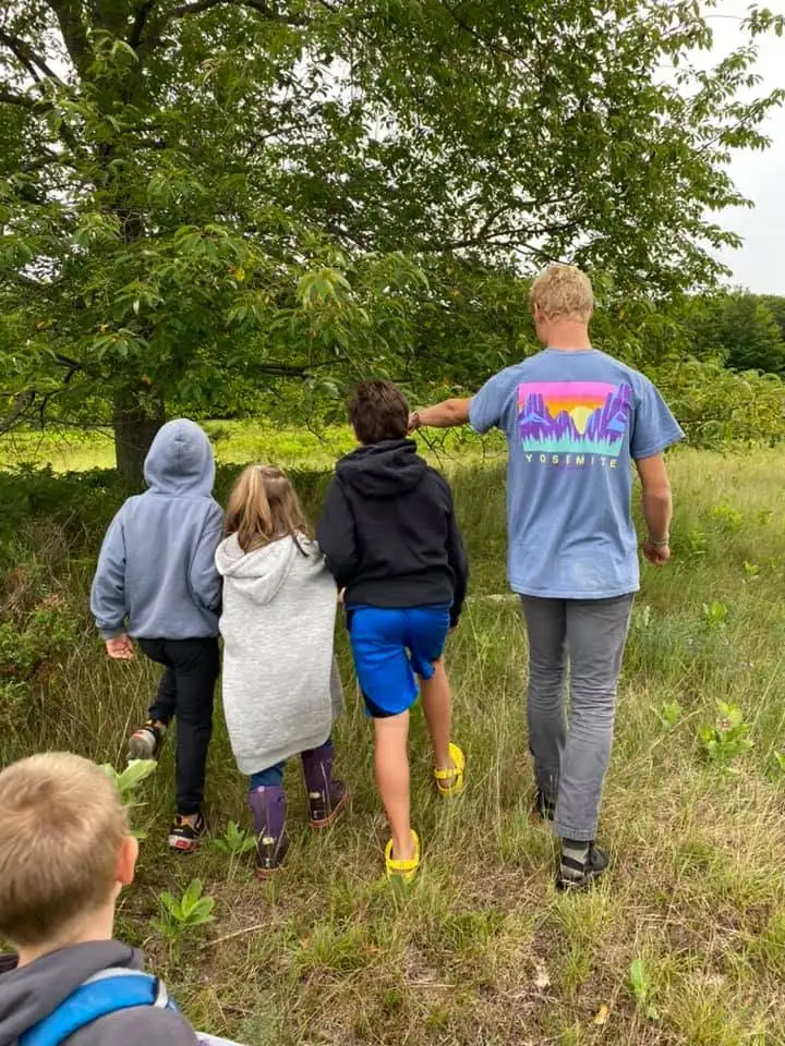 An adult and several children walk through tall grass beneath leafy trees.