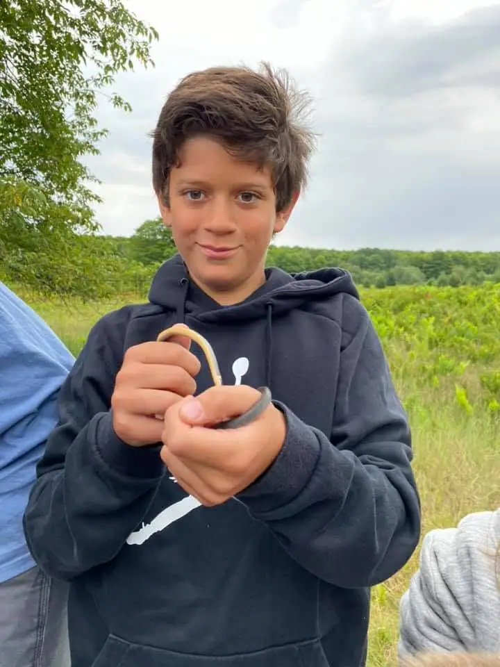 A boy in a dark hoodie holds a small curved object outdoors, with fields and trees behind him.