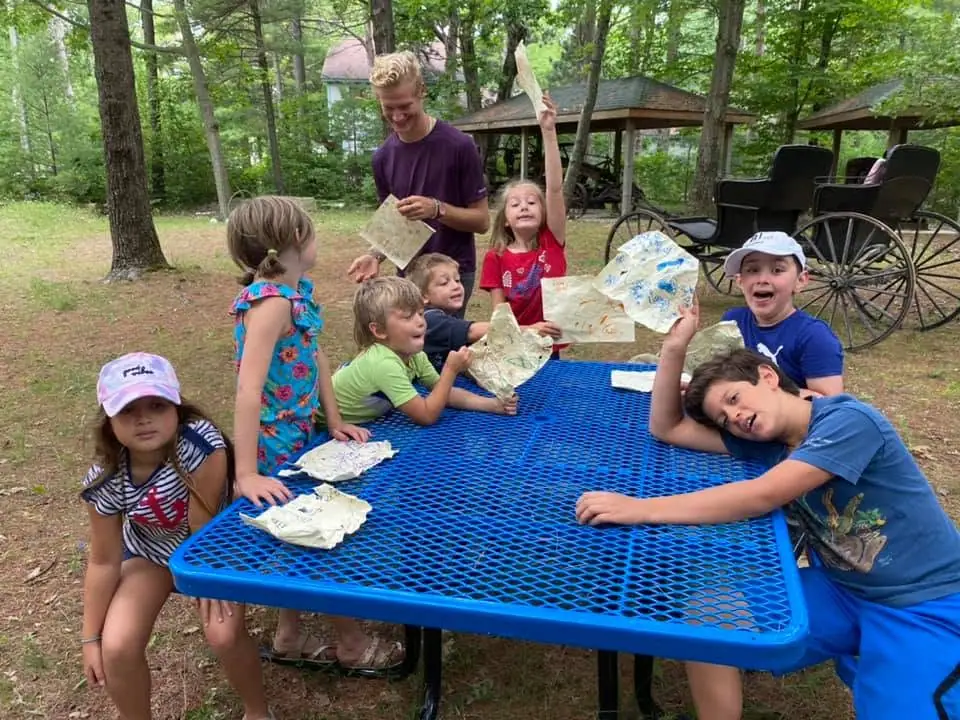 Children and an adult display papers around a blue picnic table beside a horse-drawn carriage in a wooded setting.