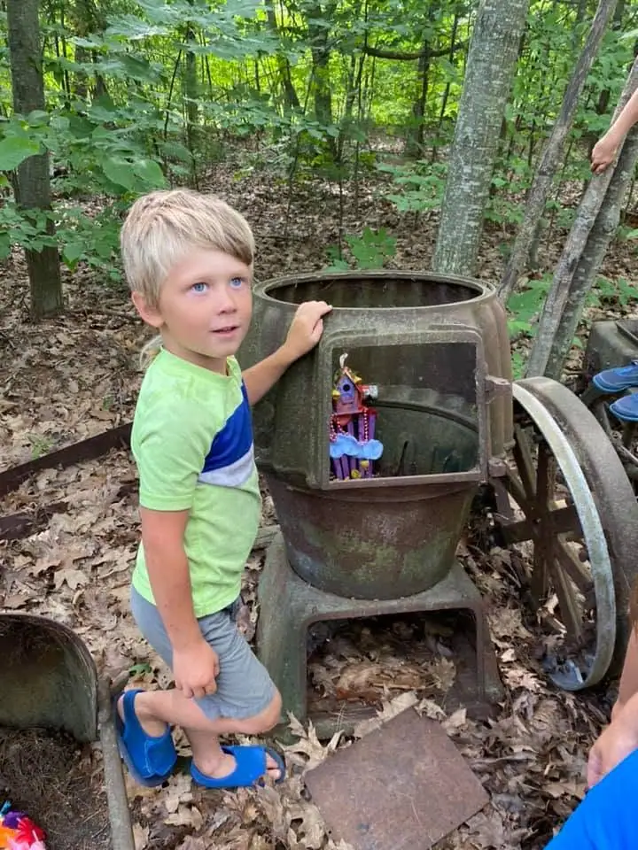 A child stands beside an old metal apparatus in a leafy wooded setting.