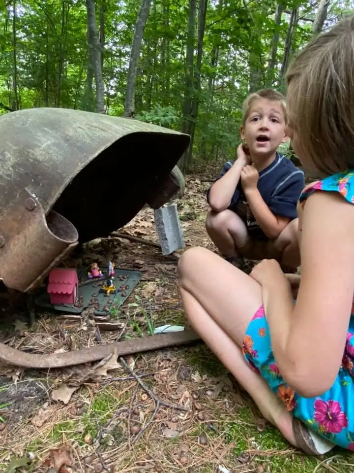 Two children examine a rusted metal object and a small toy house in a wooded area.