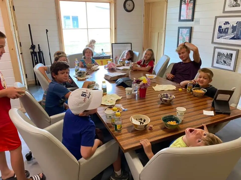 Children and adults gather around a wooden table with snacks and drinks inside a bright paneled room.