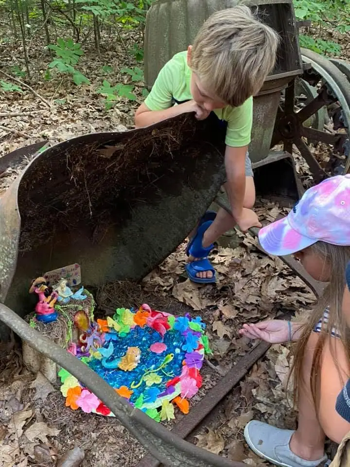 Two children examine a colorful miniature fairy garden arranged inside an old wheelbarrow in the woods.
