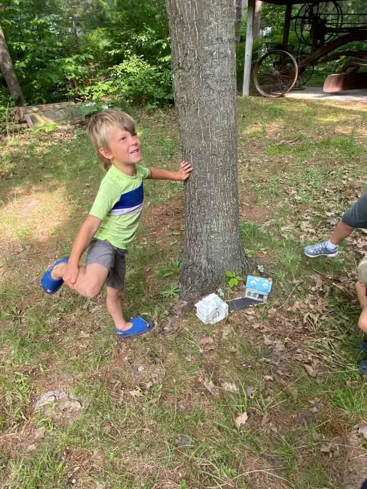 Child smiling beside a tree while holding up one leg in a grassy, wooded yard.