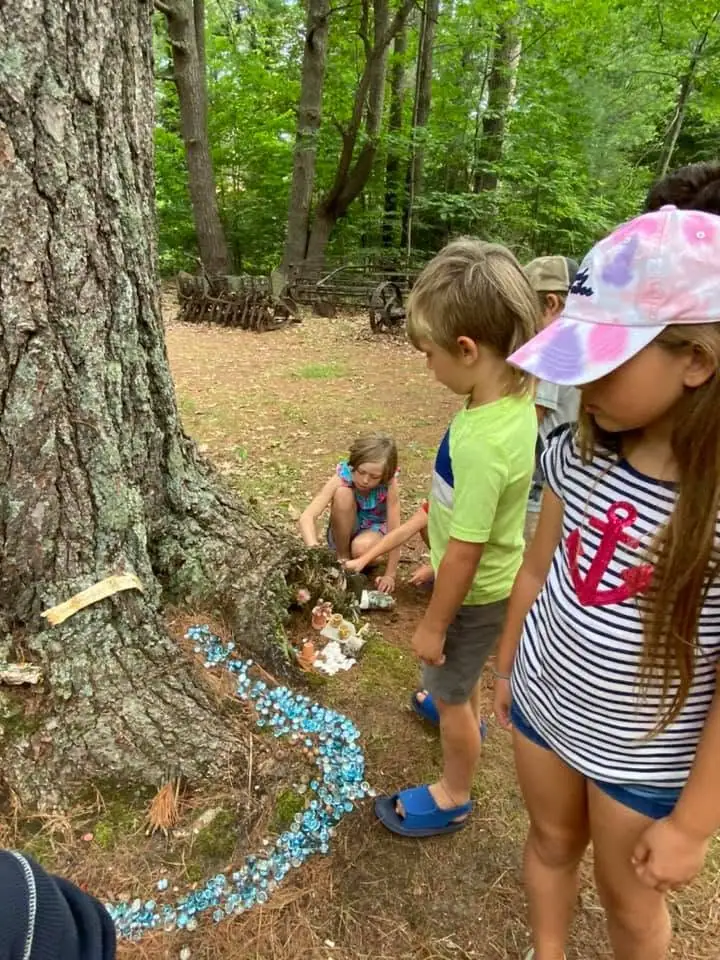 Children gather around a large tree and a winding arrangement of blue beads and miniature decorations in the woods.