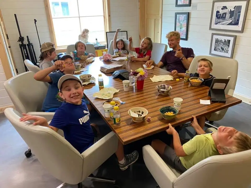 Children and an adult gather around a large wooden table with food and drinks in a bright room.