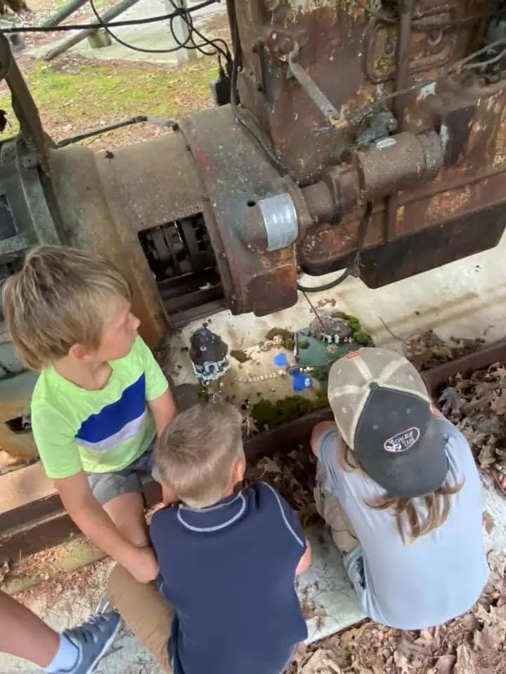 Three children examine a miniature scene beside large, rusted industrial machinery.