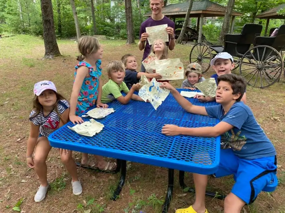 Children and an adult hold illustrated maps around a blue picnic table beneath trees, with a carriage in the background.