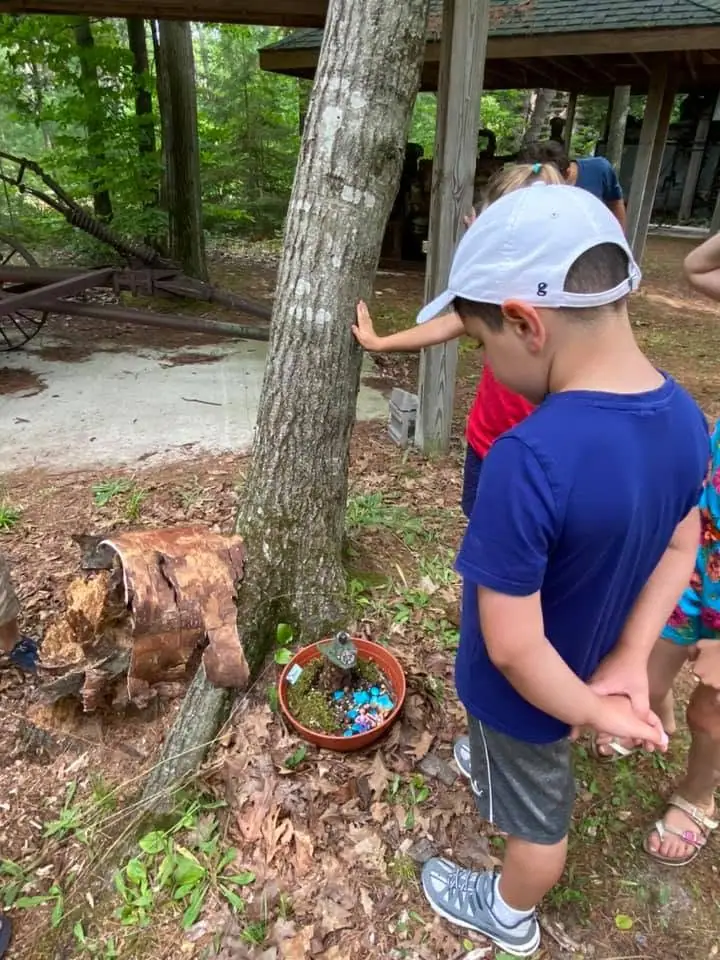 Children gather beside a tree, a colorful planter, and rusted equipment in a wooded outdoor area.