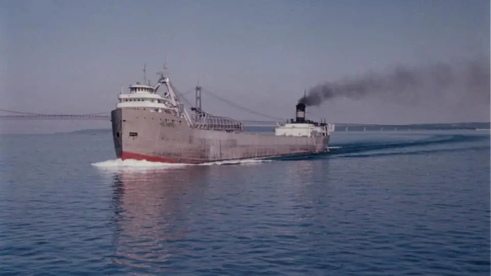 A large freighter travels across the water beneath a long suspension bridge, trailing dark smoke.