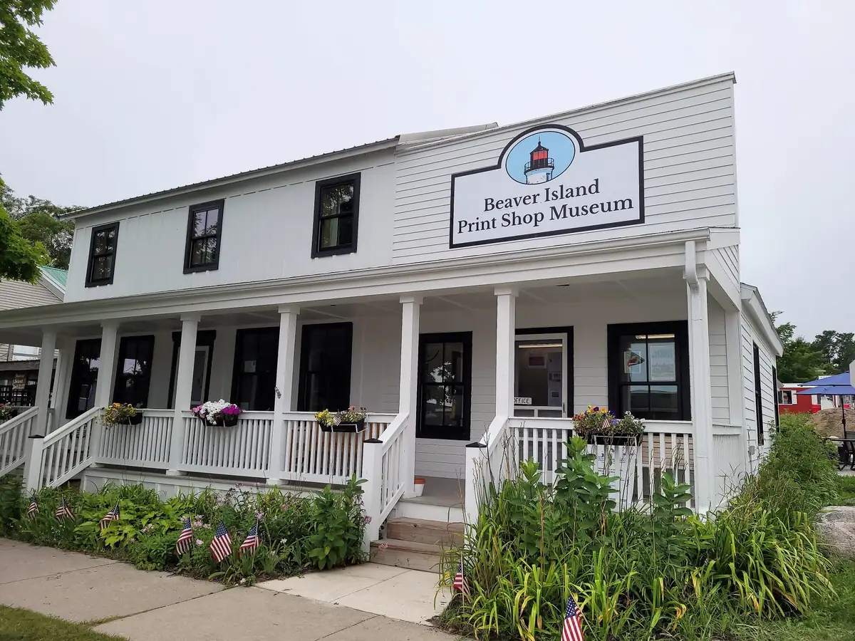 White two-story museum building with a covered porch, flower boxes, flags, and a large lighthouse sign.