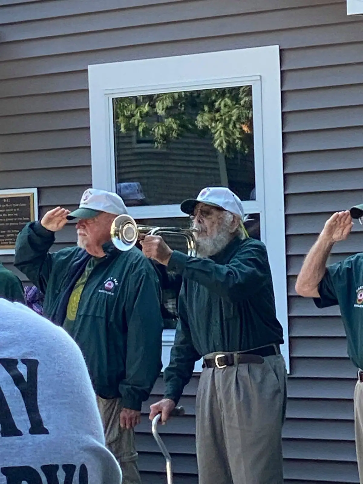 Three men in matching jackets and caps stand outside as one plays trumpet and two raise saluting hands.