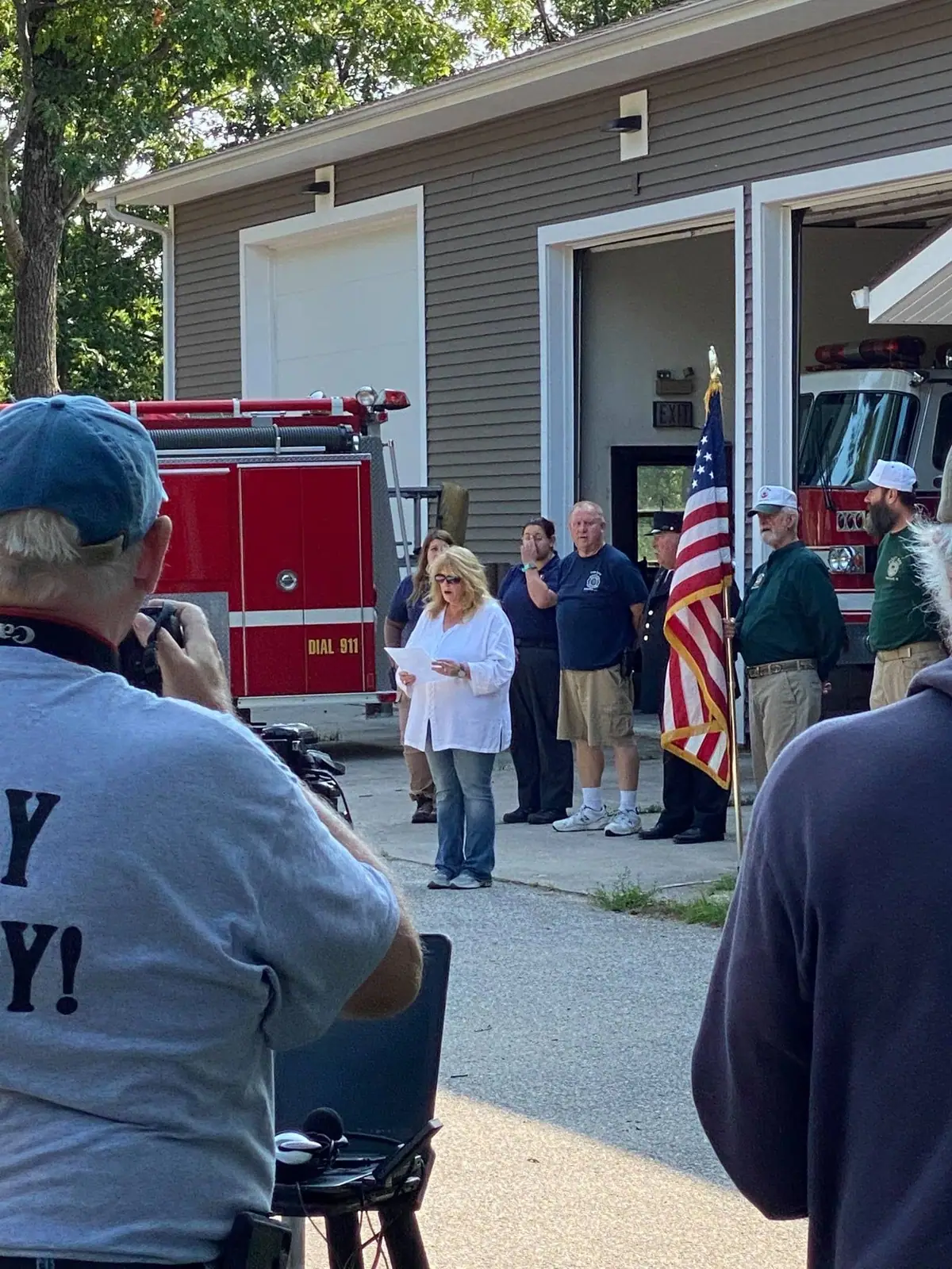 People gather outside a fire station as a woman reads from a paper beside a red fire truck and American flag.