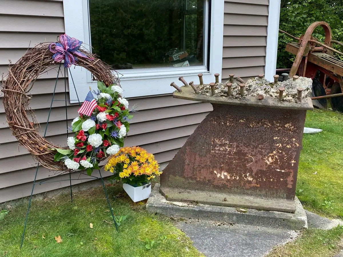 A flower-decorated wreath and potted chrysanthemums stand beside a rusted metal object outside a house.