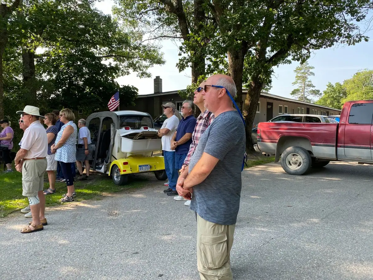 Adults stand in a line beside a paved street, with a utility cart, pickup truck, flag, trees, and a building behind them.