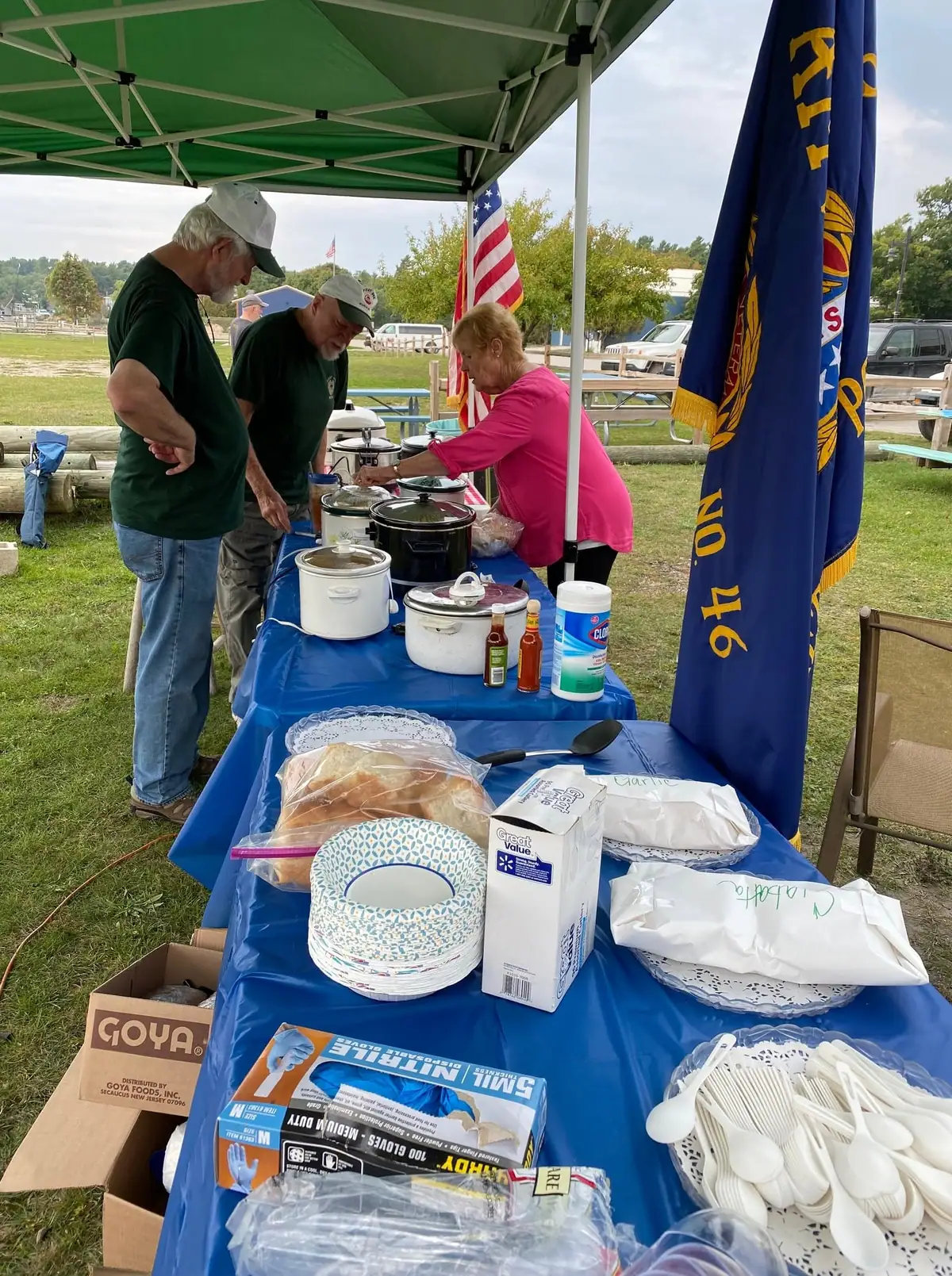 Three adults serve food from slow cookers at a table beneath a green canopy.