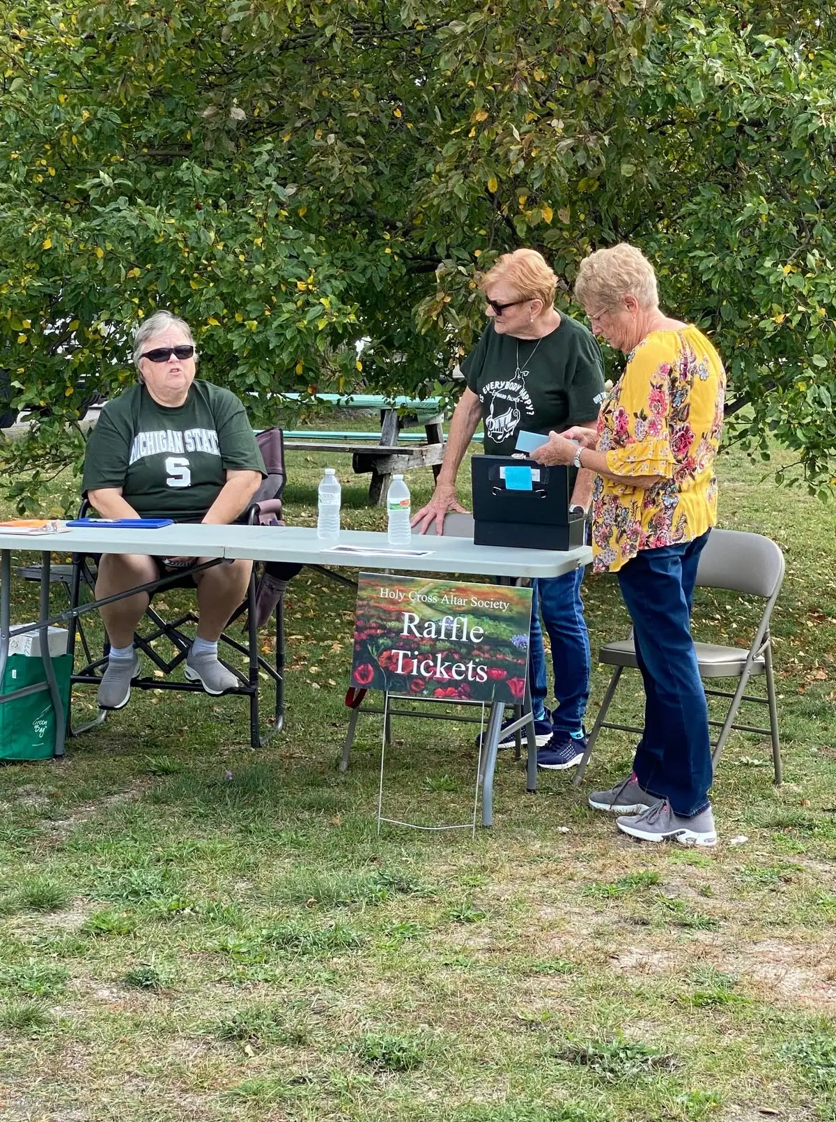 Three women operate an outdoor raffle table beneath leafy trees, with a sign advertising raffle tickets.