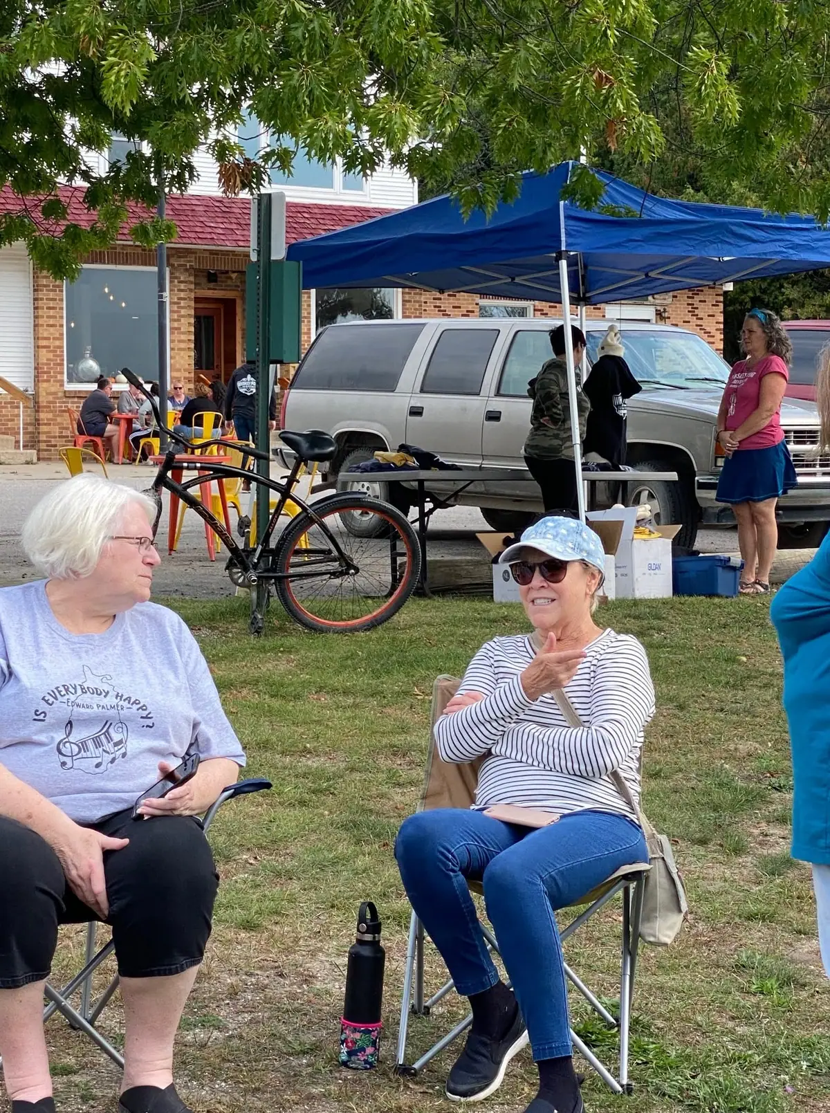 Two women sit in folding chairs beneath a blue canopy beside a bicycle and parked vehicle.