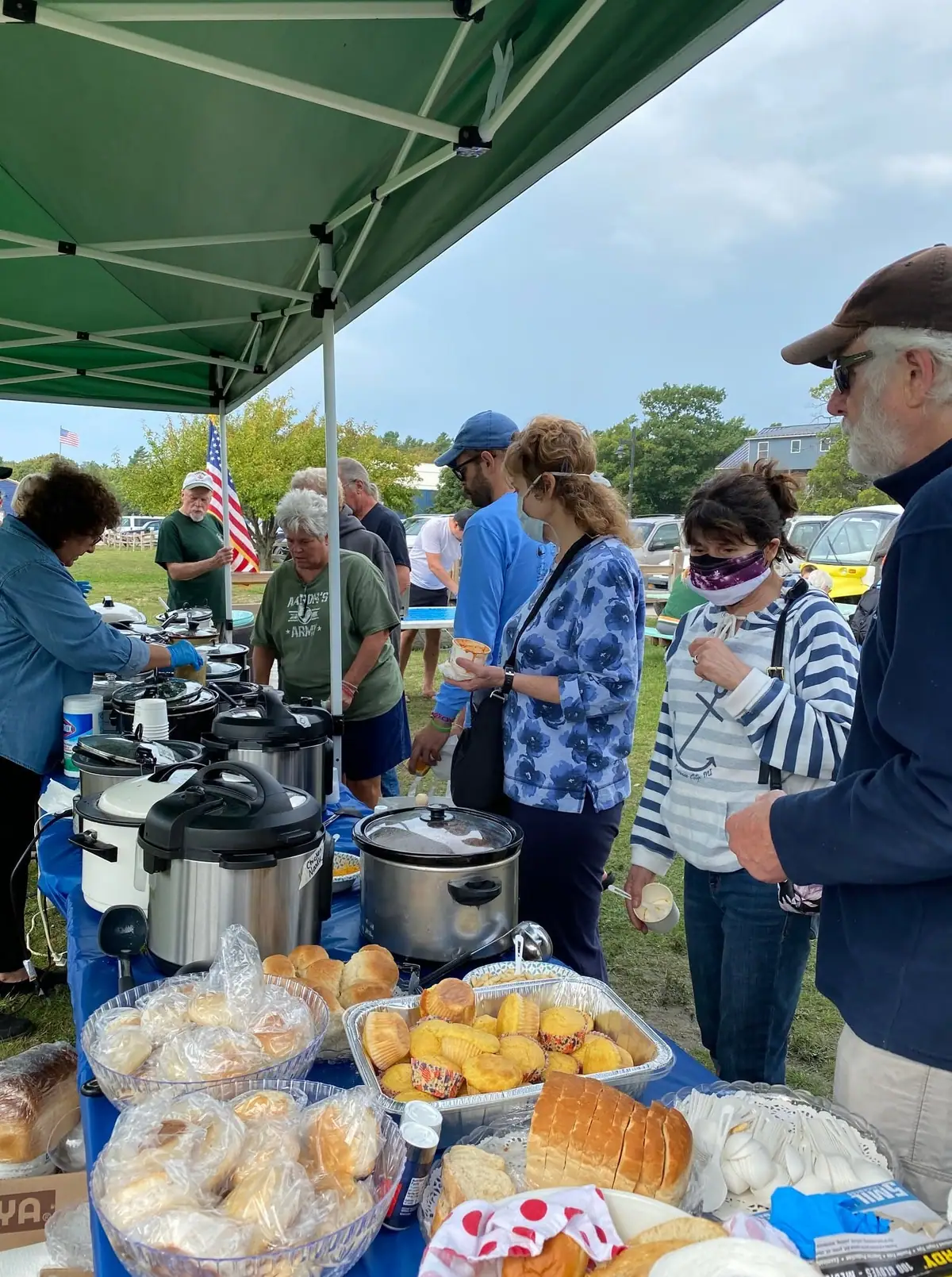Adults serve food from a table of pots, bread, rolls, and muffins beneath a green canopy.