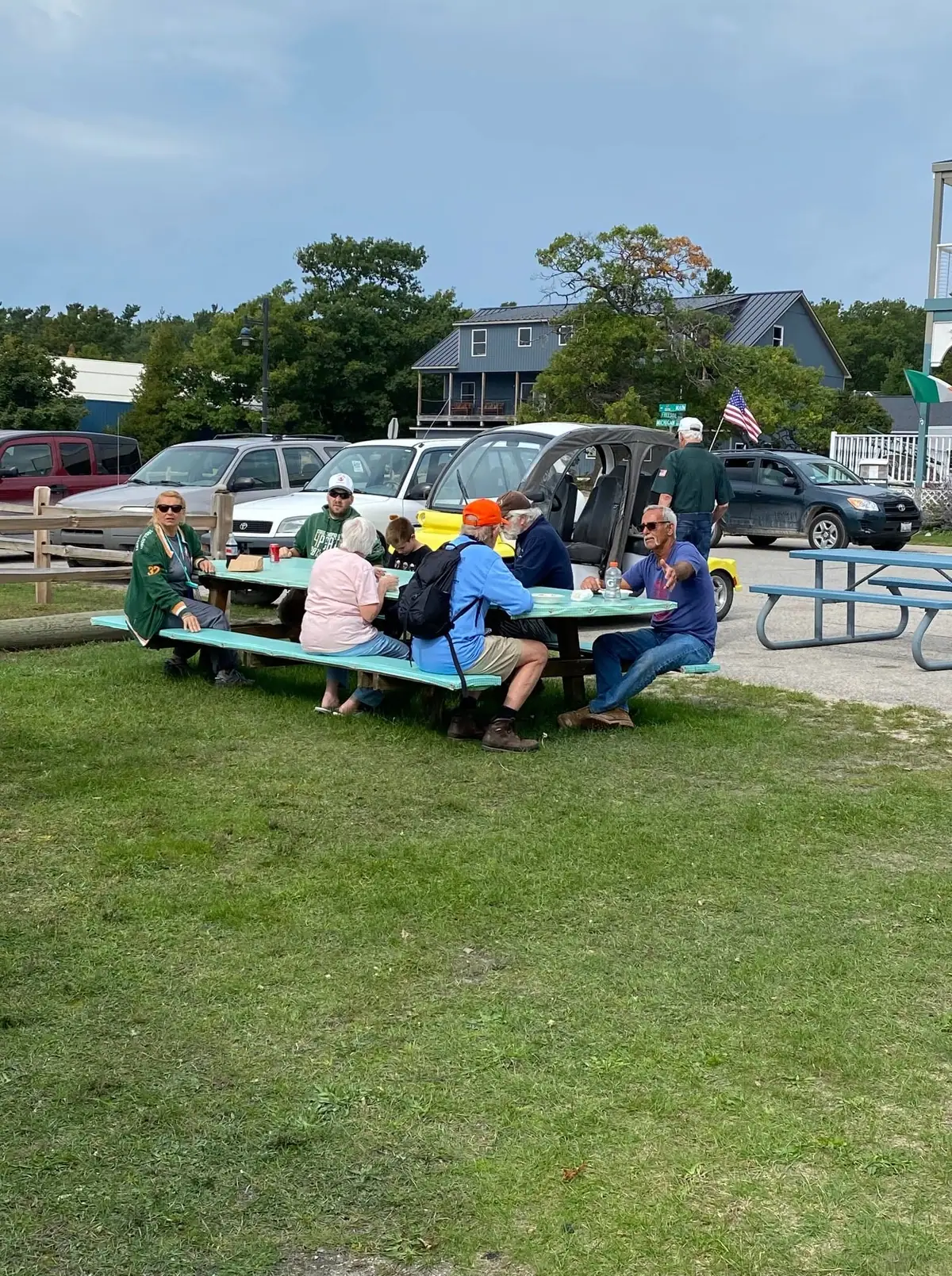 People gather around a picnic table on a grassy lawn beside parked cars and buildings.