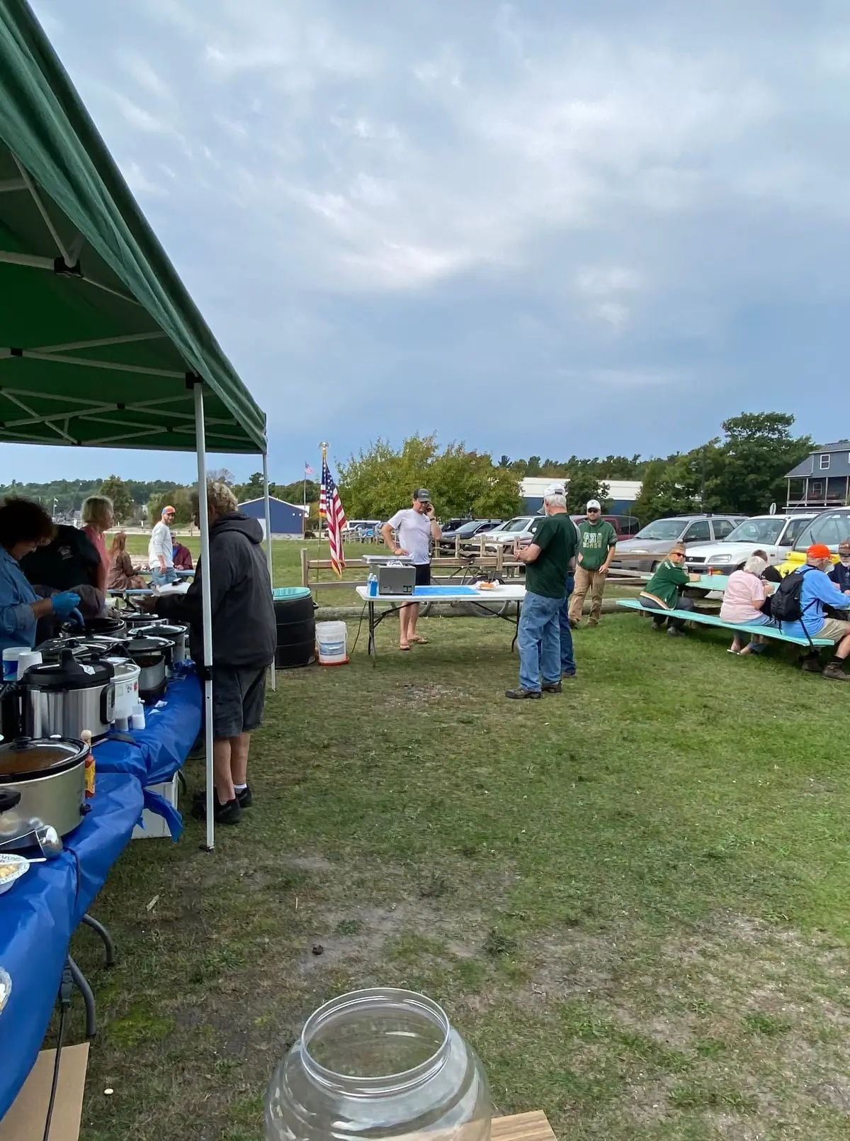 People gather outdoors around picnic tables, a food-serving tent, and an American flag.