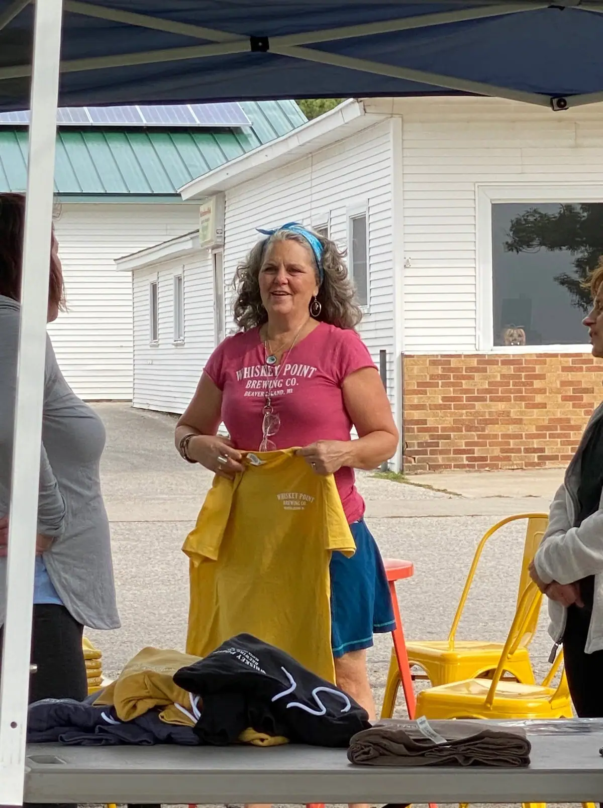 A woman displays a yellow T-shirt beside a table of folded clothing beneath an outdoor canopy.