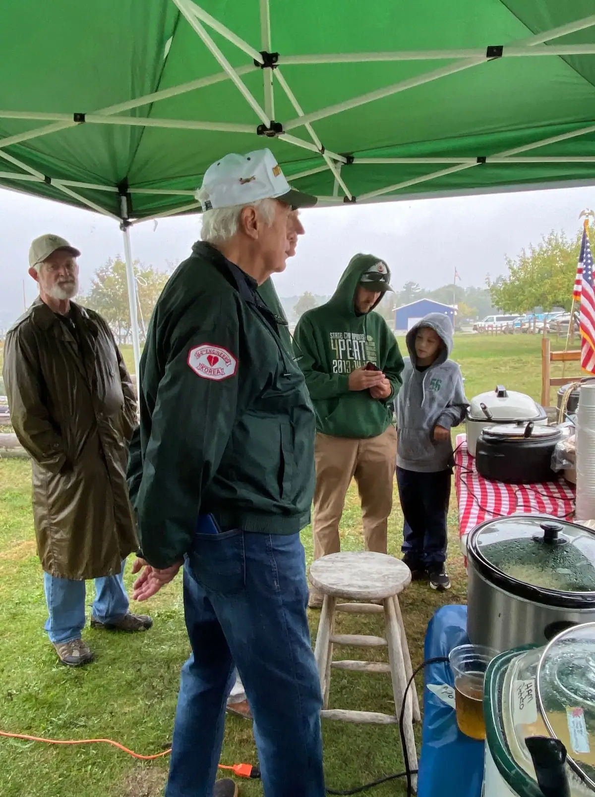 People gather beneath a green canopy beside a table of covered pots, drinks, and serving supplies.
