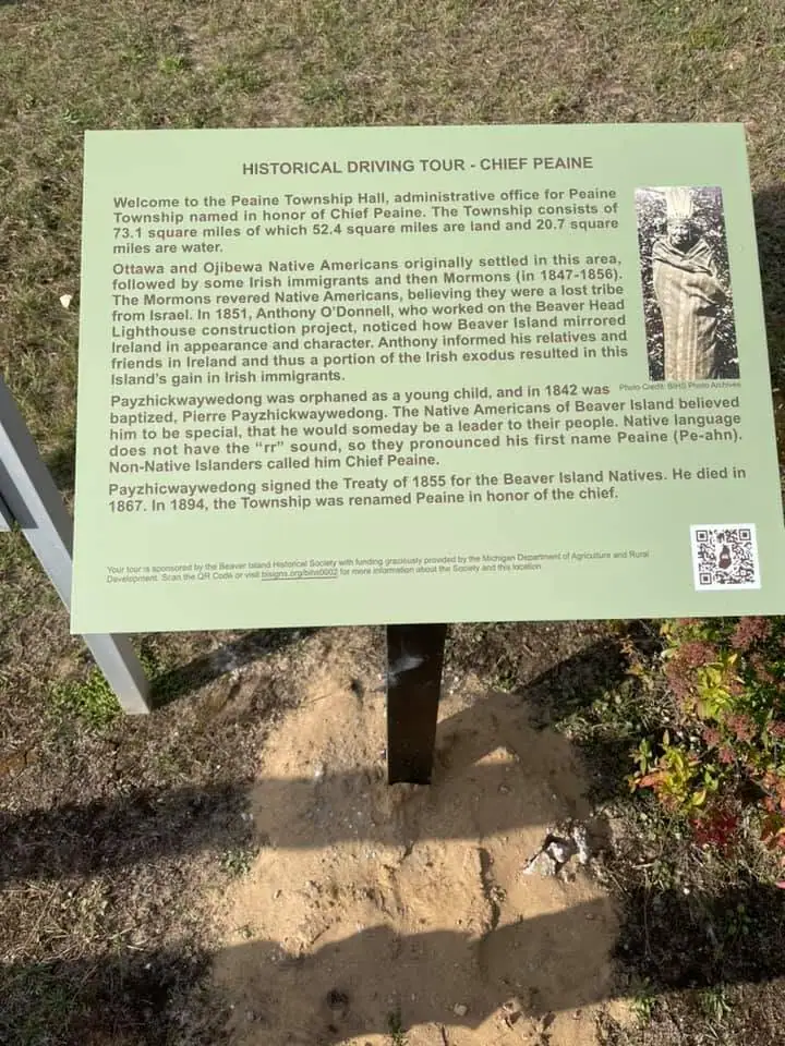 A green historical interpretive sign stands outdoors beside a small portrait and QR code.