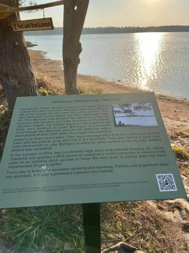 An interpretive sign and directional sign stand beside a wooded shoreline with sunlight reflecting on the water.