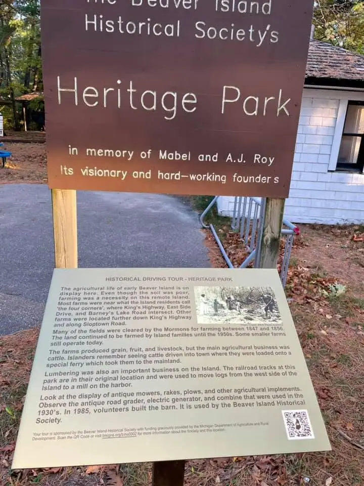 A wooden heritage sign and an agricultural-history information panel stand beside a paved path and white building.