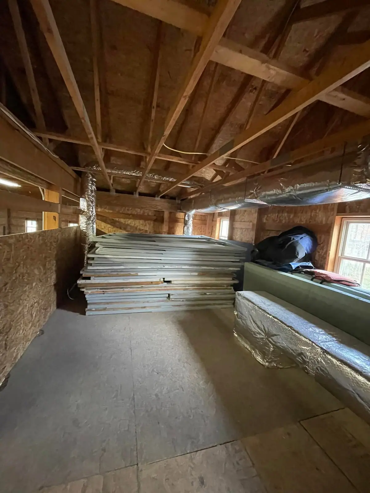 Interior storage space with exposed rafters, ductwork, windows, and a large stack of framed plywood panels.