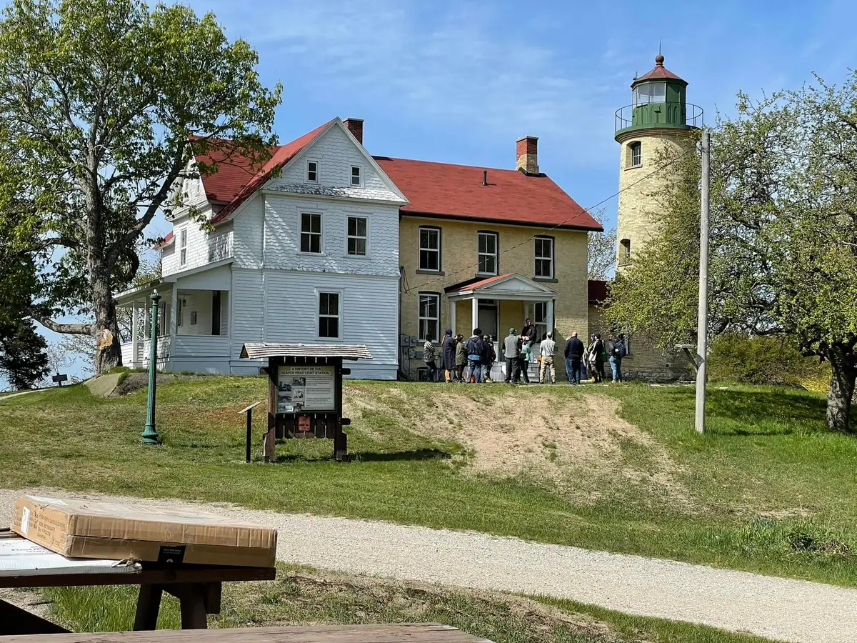 A group stands beside a white building and stone lighthouse tower, with an interpretive sign and picnic table in front.
