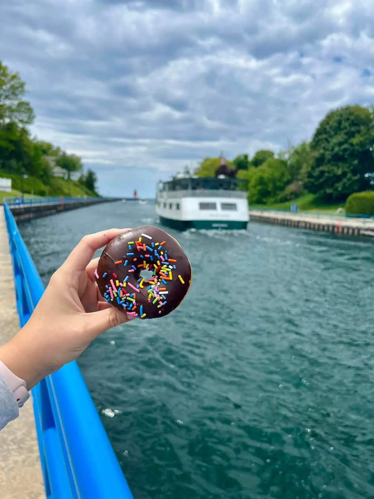 A hand holds a chocolate-glazed sprinkle donut in front of a ferry traveling along a tree-lined waterway.