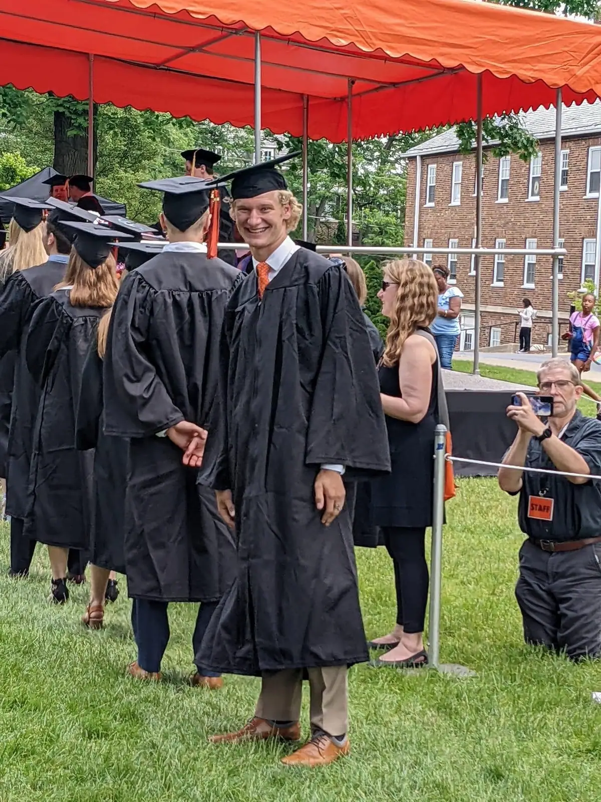 A smiling graduate in academic regalia stands beneath a red canopy while other graduates and a photographer surround him.
