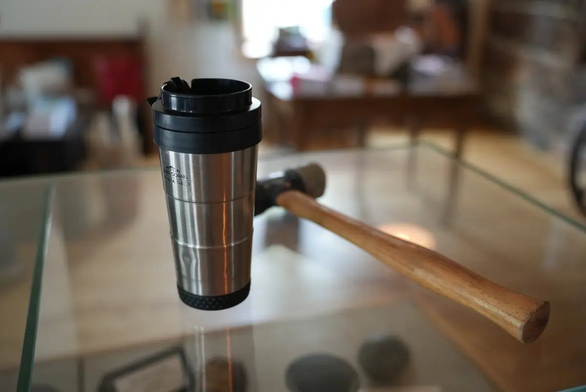 A stainless-steel travel mug and wooden mallet rest on a glass display case in an exhibit space.