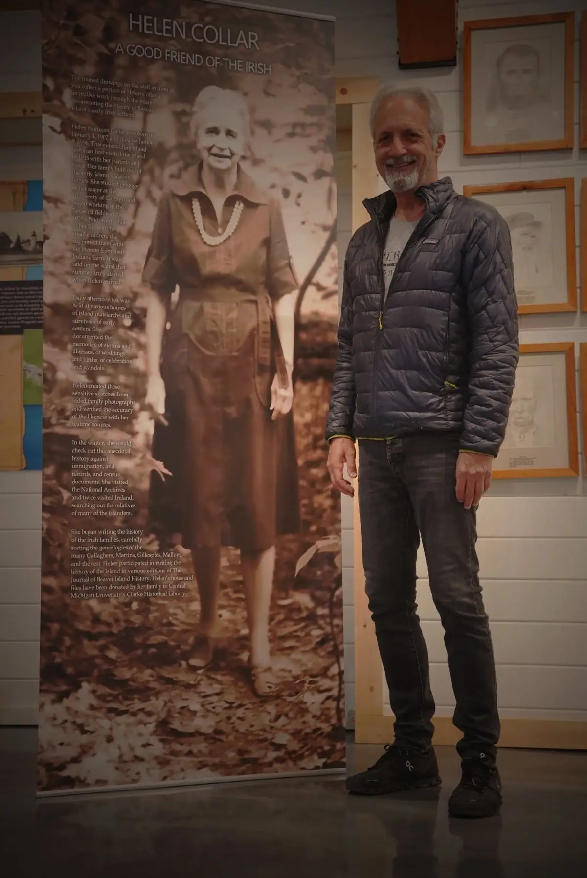 A man stands beside a sepia-toned exhibition banner and framed drawings inside a gallery.