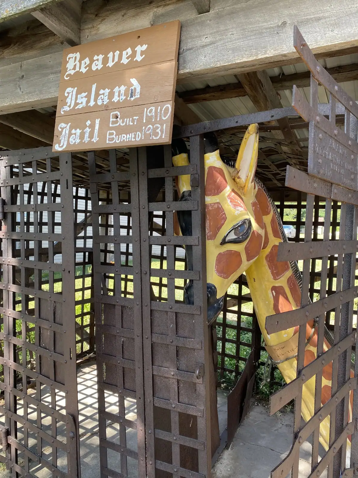 A painted giraffe sculpture stands behind metal bars beneath a wooden jail sign.