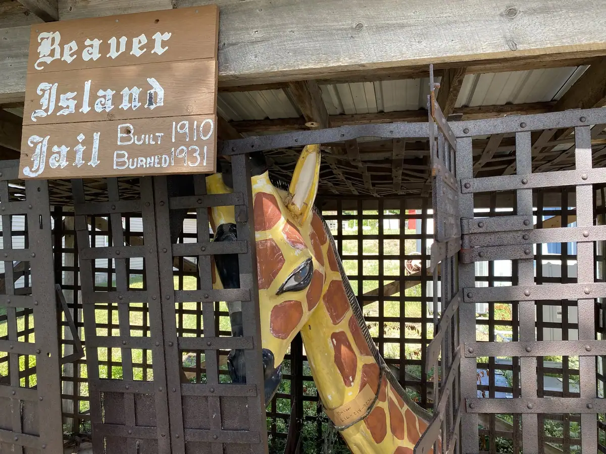 A painted giraffe sculpture stands behind a metal cage beneath a wooden sign.