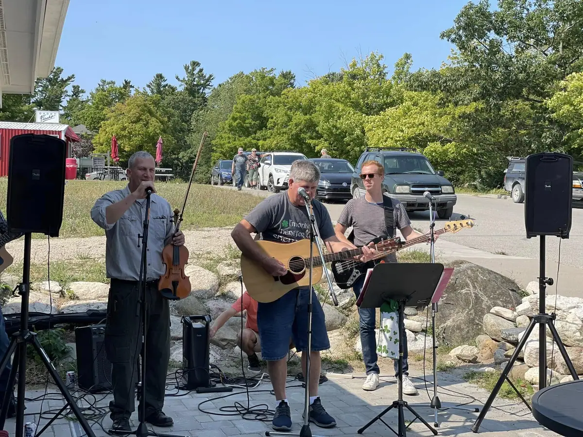 Three musicians perform outdoors with a violin, acoustic guitar, bass guitar, microphones, and speakers.