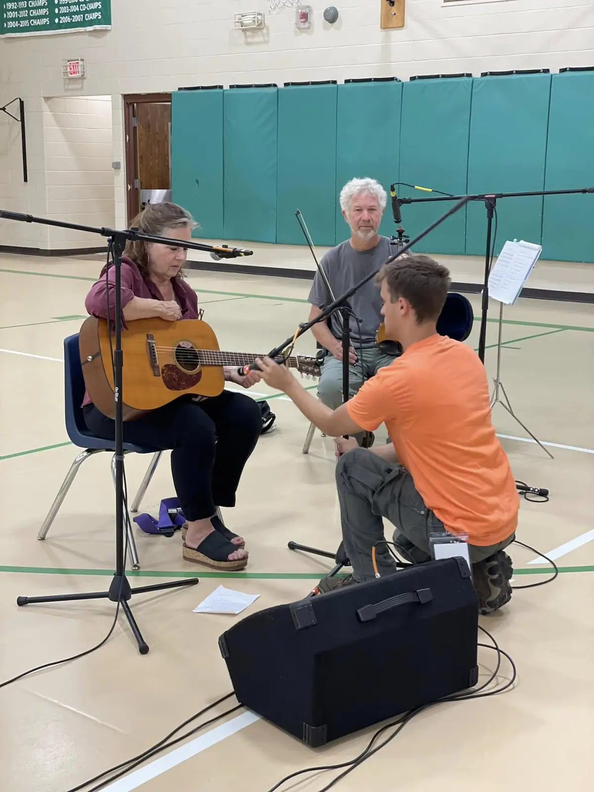 Three people rehearse in a gymnasium as a seated woman plays acoustic guitar among microphones and audio equipment.