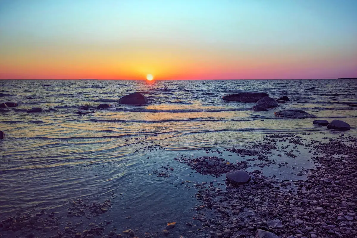 Sunset over calm water and a rocky, pebble-covered shoreline.