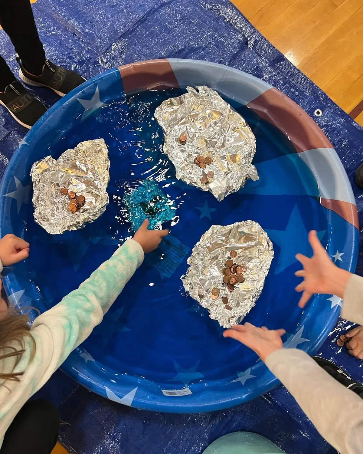 Children’s hands arrange coins on foil-covered floating shapes in a small water-filled pool.