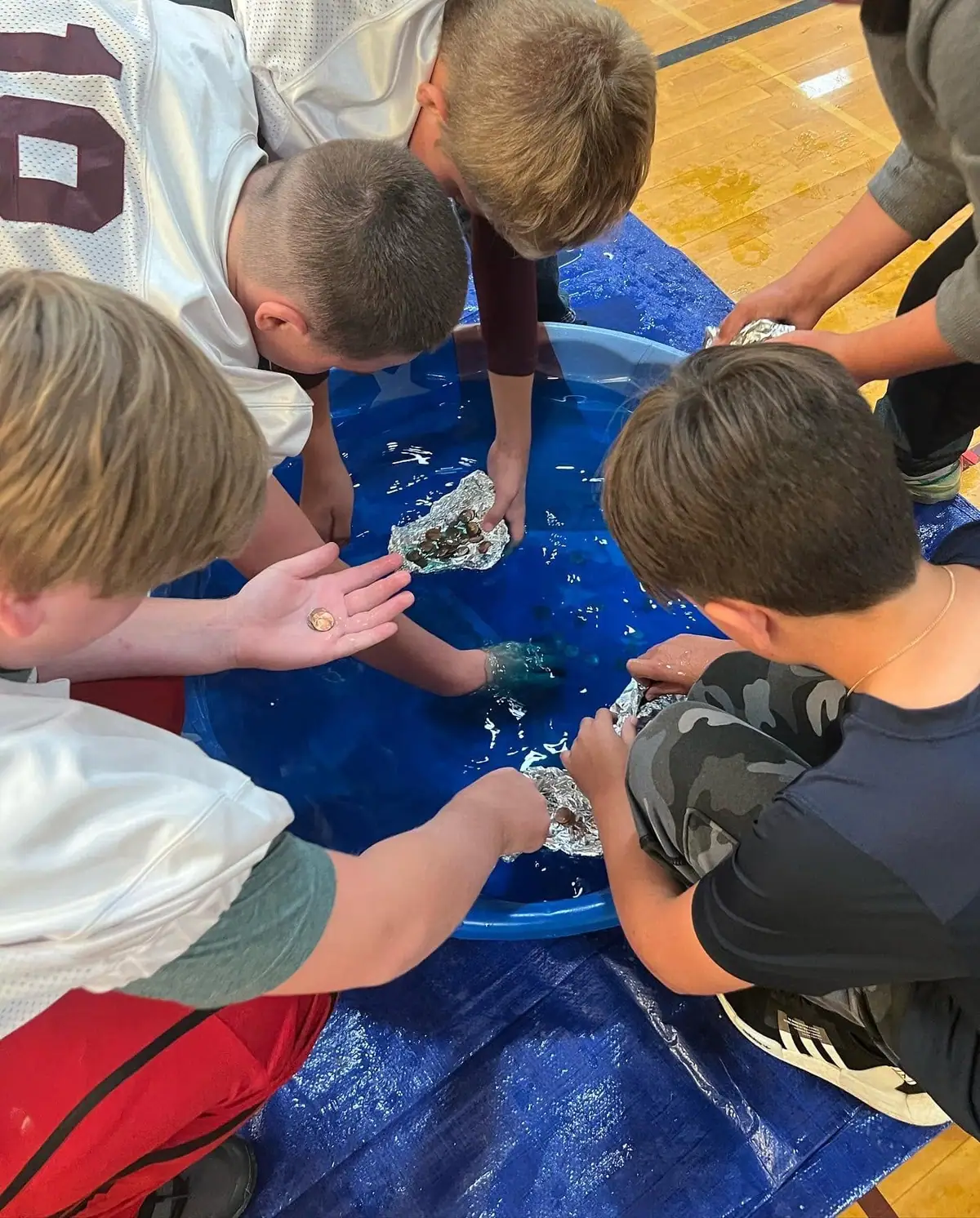 Children and an adult handle foil boats and coins in a blue water-filled tub.