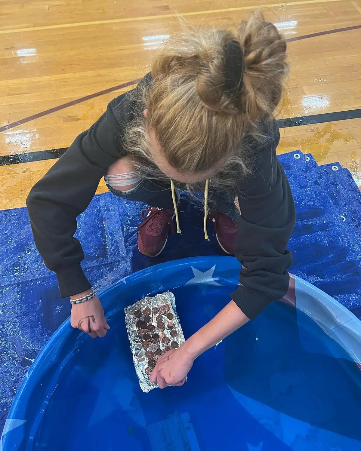 A person kneels over a blue inflatable pool while holding a foil-wrapped block of coins in the water.